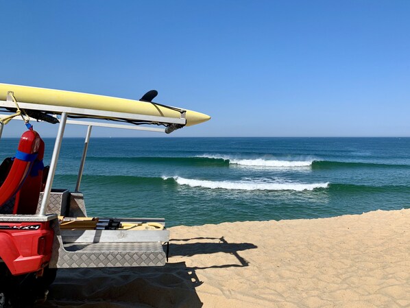 Une scène de plage avec une planche de surf posée sur un support jaune, une bouée de sauvetage rouge et un véhicule doté d’une calandre métallique.