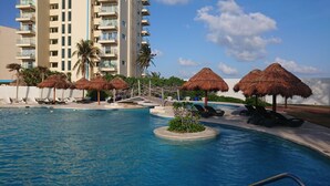 A resort pool area with lounge chairs, thatched umbrellas, and a palm tree. - 2 Steps to the Beach 2 bedroom apartment (Zona Hotelera)