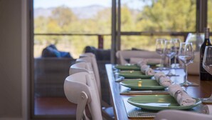 Mesa de comedor servida con platos verdes, servilletas blancas y copas de vino, con vistas a árboles y montañas a través de amplios ventanales.