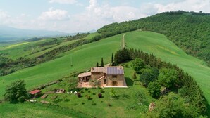 Une maison en pierre surmontée d’un toit équipé de panneaux solaires, entourée de champs verdoyants et d’arbres, avec en arrière-plan une vue sur des collines ondoyantes.