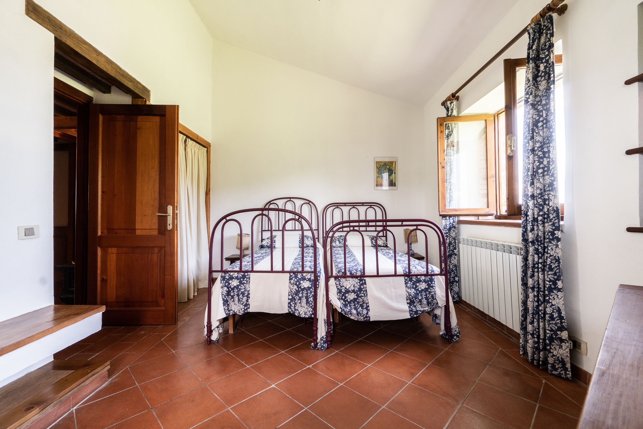 Una habitación con dos literas metálicas, una puerta de madera, un radiador y una ventana con cortinas.