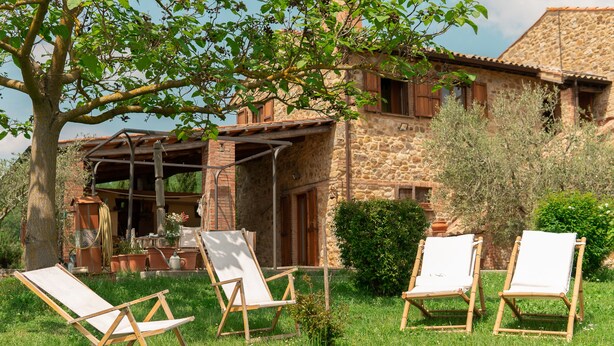 Un bâtiment en pierre à l’aspect rustique, doté d’un espace extérieur abrité avec des chaises en bois, et d’une pelouse verdoyante et luxuriante.