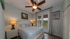 A bedroom with a bed, two bedside tables with lamps, a ceiling fan, a door with a view, and a painting on the wall. - Gator Cove: Quaint 2-bedroom, 1-bath cottage, sleeps 4. Great for a peaceful retreat. (Oviedo)