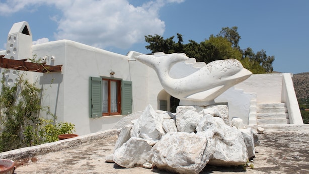 Un edificio bianco con una scultura particolare sul tetto, una finestra con persiane verdi e un cielo azzurro intenso.