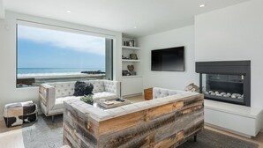 Un salon moderne doté d’une grande fenêtre donnant sur la plage, d’une cheminée et d’un téléviseur à écran plat fixé au mur.