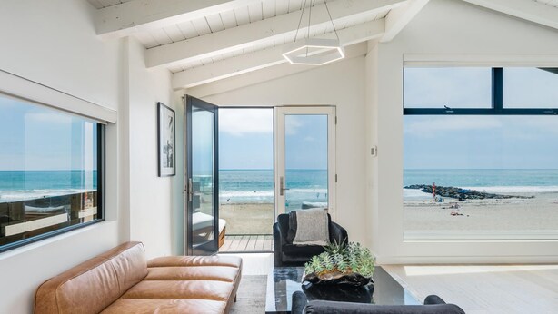 Un salon moderne avec un canapé en cuir, un fauteuil noir et une table basse agrémentée d’une plante. De grandes portes vitrées donnent accès à un balcon offrant une vue sur la plage.