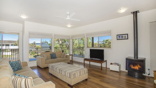 A spacious living room with a sofa, ottoman, and a television. A fireplace is visible, and there are large windows offering a view of the outdoors. - Mitchellmook in Mollymook (Mollymook Beach)