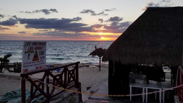Escena de playa con atardecer, una cabaña con techo de paja y un letrero de advertencia.