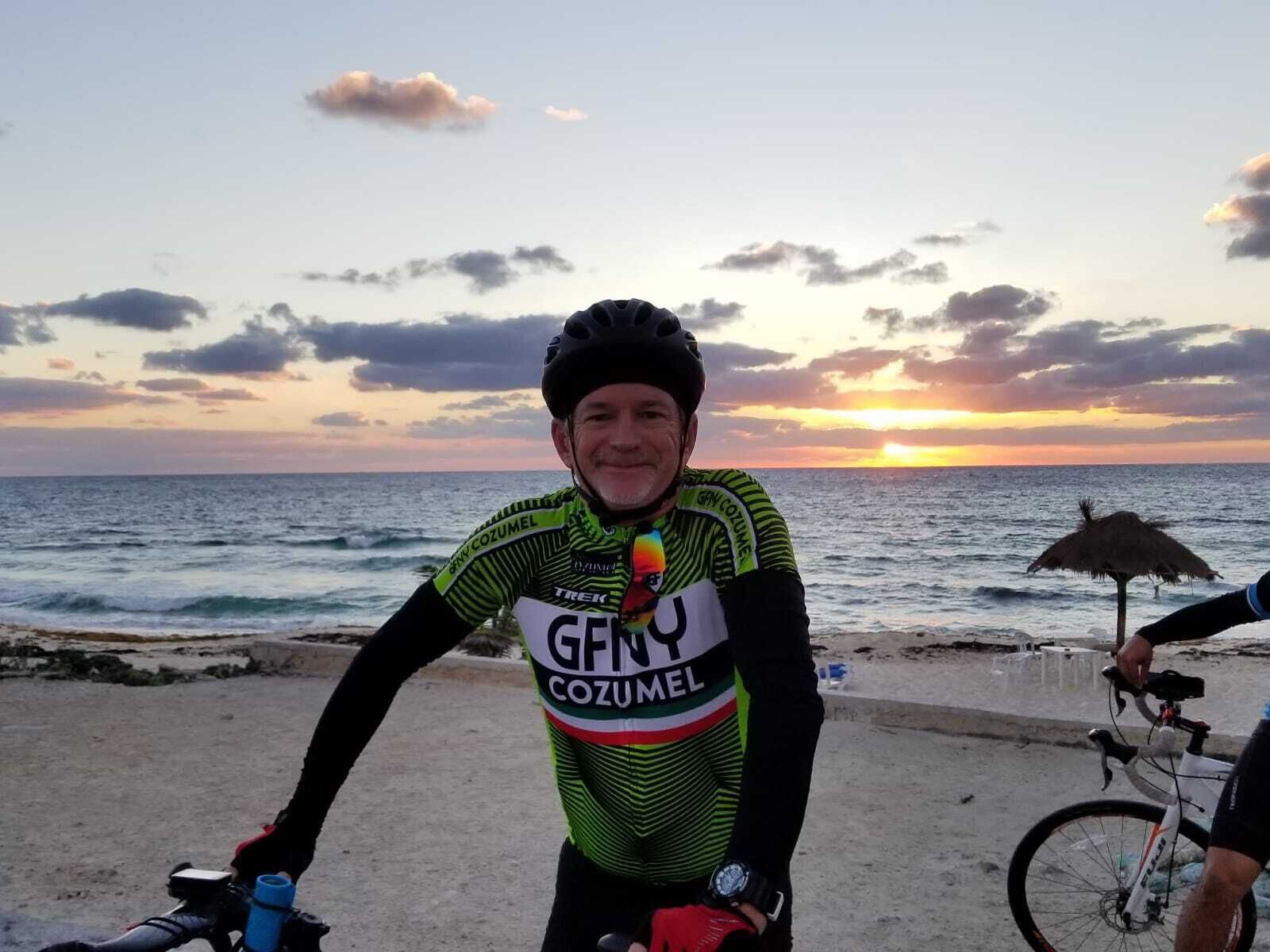 A person wearing a cycling jersey with the brand 'GENY COZUMEL' and a watch, standing by a beach with a sunset in the background.