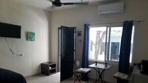 Una habitación con cama, una mesita pequeña con dos sillas, una ventana con cortinas, un ventilador de techo y un equipo de aire acondicionado.