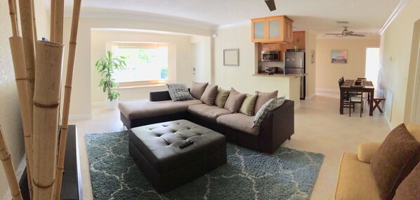 A living room with a sectional sofa, ottoman, and a dining area with a table and chairs. - Savers Paradise Vacation Home (Lauderdale-By-The-Sea)