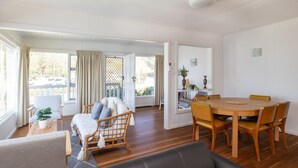 A spacious room with a dining table, chairs, a sofa, and a window with curtains. - Surf & Turf Retreat in Mollymook (Mollymook)