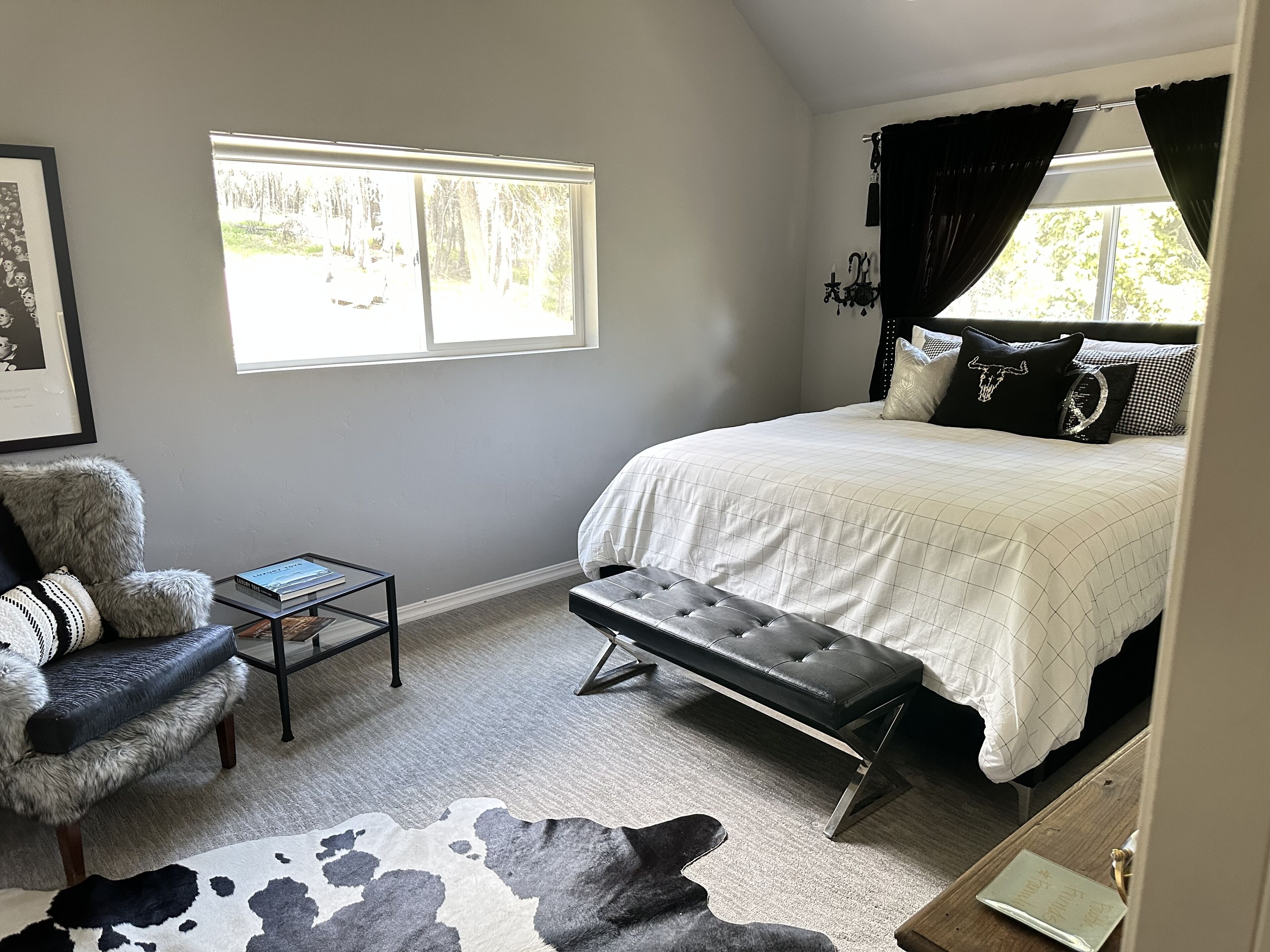A bedroom with a large bed, a bench, a chair, a small table, and a window with curtains.