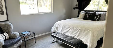A bedroom with a large bed, a bench, a chair, a small table, and a window with curtains.