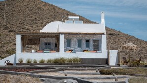A modern house with a flat roof, a small porch with colorful cushions, and a view of a desert landscape. - Le Grand Blue Villa Santorini (Santorini)