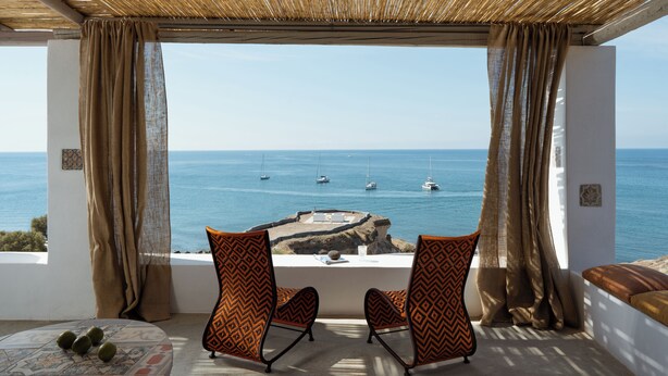 A balcony with two patterned chairs, a table with a glass, and a view of the sea with boats. - Le Grand Blue Villa Santorini (Santorini)