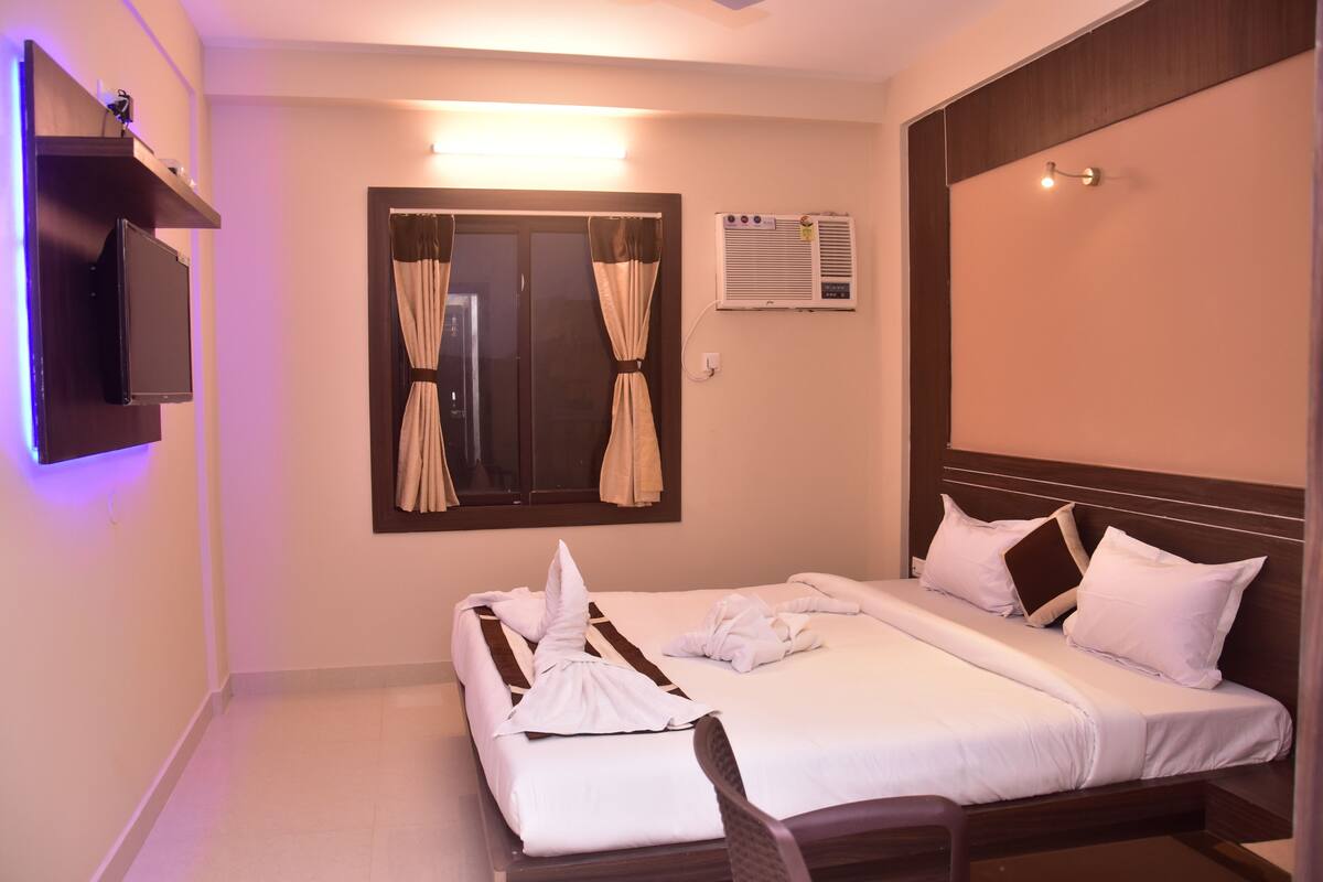 Una habitación de hotel con cama, televisor montado en la pared, unidad de aire acondicionado y ventana con cortinas.