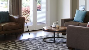 A living room with a brown leather sofa, a grey patterned rug, a round coffee table, and a view through a sliding glass door. - Cuballa - Just a few minutes flat  walk to town & the river .Modern clean house (Bright)