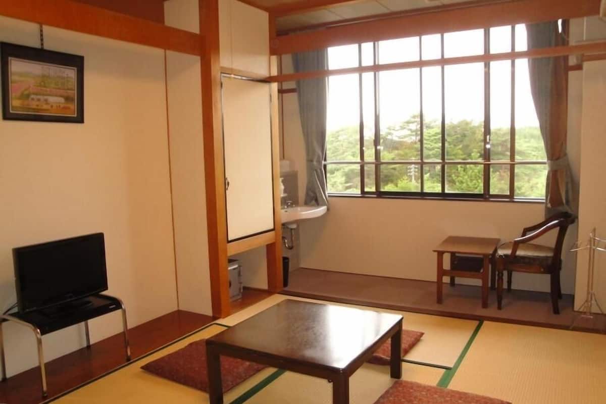 A room with tatami mats, a television, a table, a chair, and a window with a view of greenery.