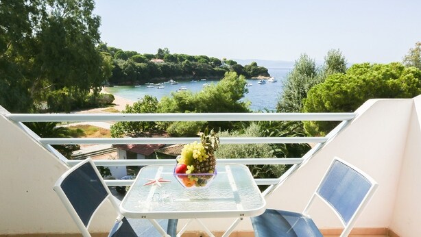 A balcony with a table and two chairs, overlooking a coastal area with boats and greenery.