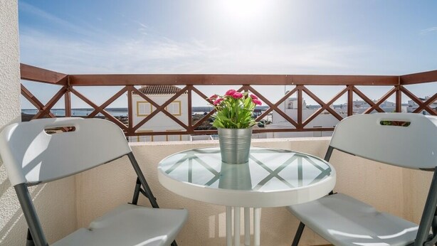 A balcony with a glass table and two white chairs, a metal pot with flowers, and a view of the cityscape.