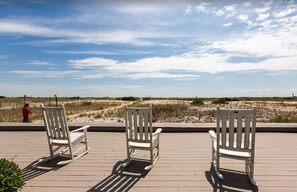 Tres sillas mecedoras en una terraza de madera con vistas a una playa con dunas de arena y cielo despejado.