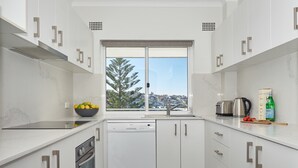 A modern kitchen with white cabinets, a dishwasher, a window with a city view, and a countertop with a kettle and a bottle. - Ocean View Apartment with a Stroll to Bronte Beach (Bronte)