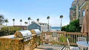 A barbecue area with stone and metal grill, a wooden fence, and a view of the beach and ocean.
