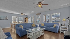 Ein Wohnzimmer mit zwei blauen Sofas, einem Glastisch, Holzböden, Deckenventilatoren und großen Fenstern mit Jalousien.