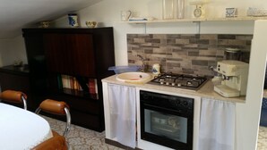 Cocina compacta con estufa a gas, horno y cafetera. Hay un lavamanos con un escurreplatos azul, un armario de madera y una zona de comedor con mesa y sillas.
