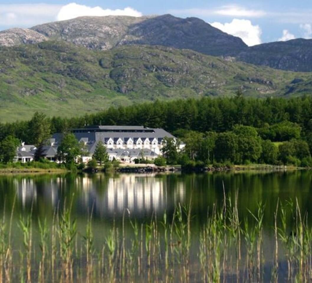A lakeside hotel with a large building featuring a symmetrical facade and multiple windows, surrounded by lush greenery and mountains in the background.