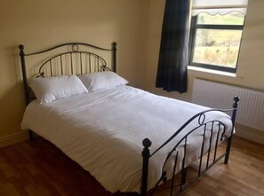 A single bed with a metal frame and white bedding in a room with wooden flooring and a window with a view of greenery.