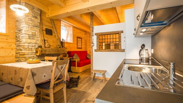 Een gezellig chaletinterieur met een stenen open haard, een houten eettafel, een rode bank en een keuken met een spoelbak en fornuis.