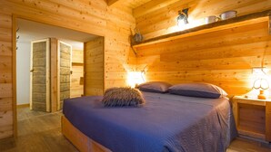 Een knus slaapkamer van een houten blokhut, met een bed, kussens, een nachtkastje en een lamp.