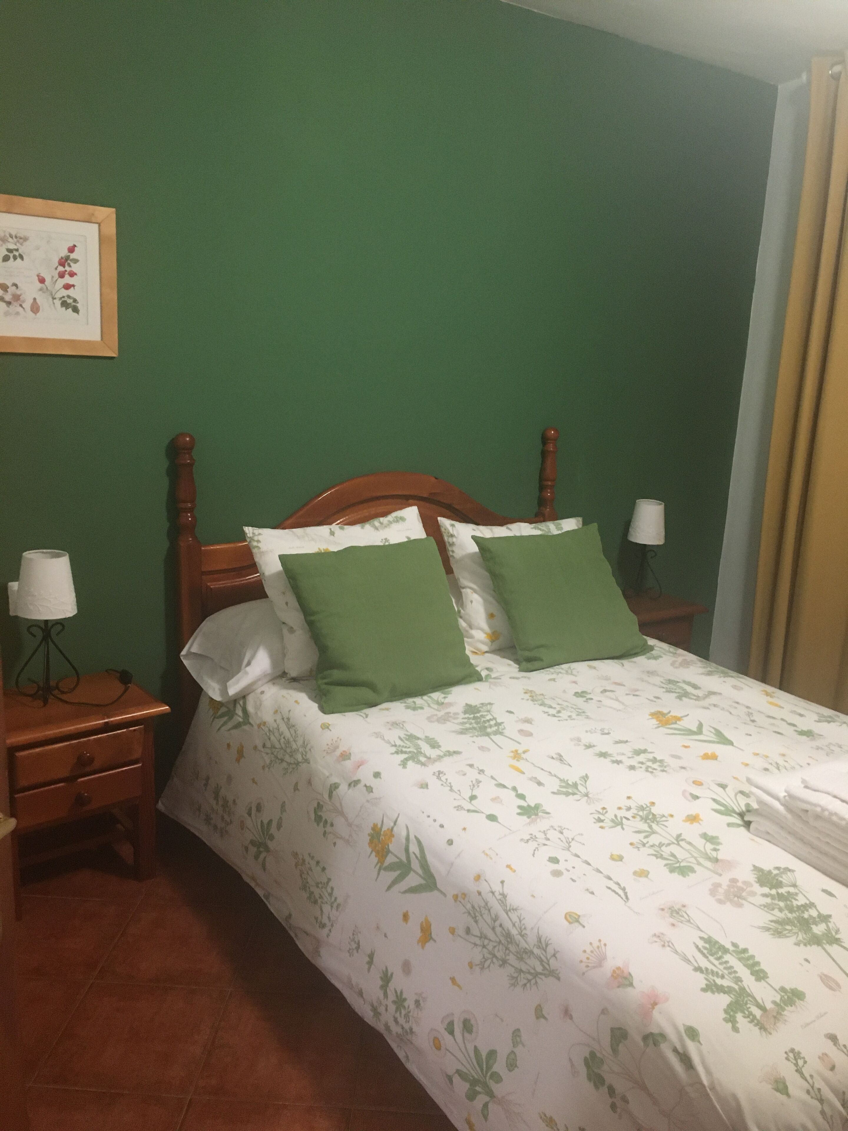 A bedroom with a wooden bed, green pillows, and a floral bedspread. There is a nightstand with a lamp and a framed picture on the wall.