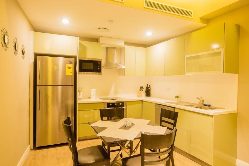 Cocina compacta con refrigerador de acero inoxidable, microondas y estufa. Hay una mesa de comedor con sillas y una pequeña isla con un florero.
