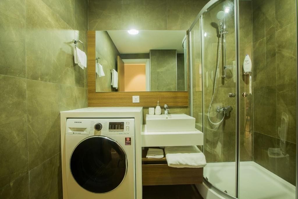 Ett litet hotellbadrum med tvättmaskin, handfat, spegel och en dusch med glasväggar.