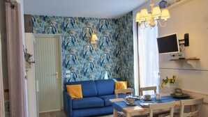 A living room with a blue sofa, a dining table with chairs, a TV mounted on the wall, and a patterned wallpaper. - Charming apartment, mit pools, in a residence  just a few minutes from the lake (LAZISE)