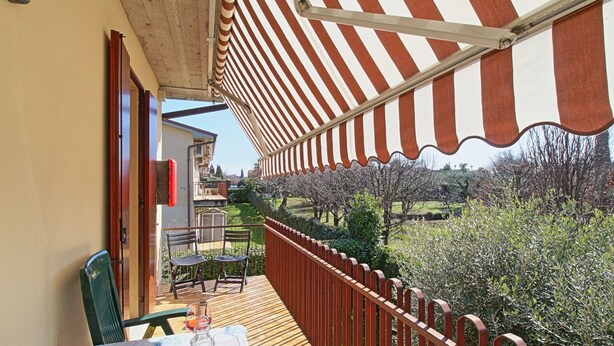A balcony with a striped awning, a table with chairs, and a view of a garden with trees and a building in the background. - Charming apartment, mit pools, in a residence  just a few minutes from the lake (LAZISE)