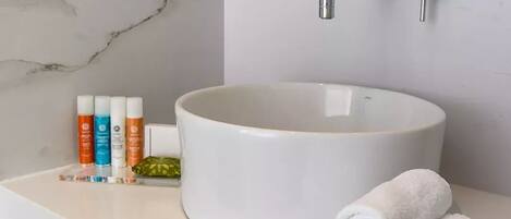 A bathroom with a marble wall, a round sink, and a mirror. There are toiletries on the counter and towels stacked on the side.