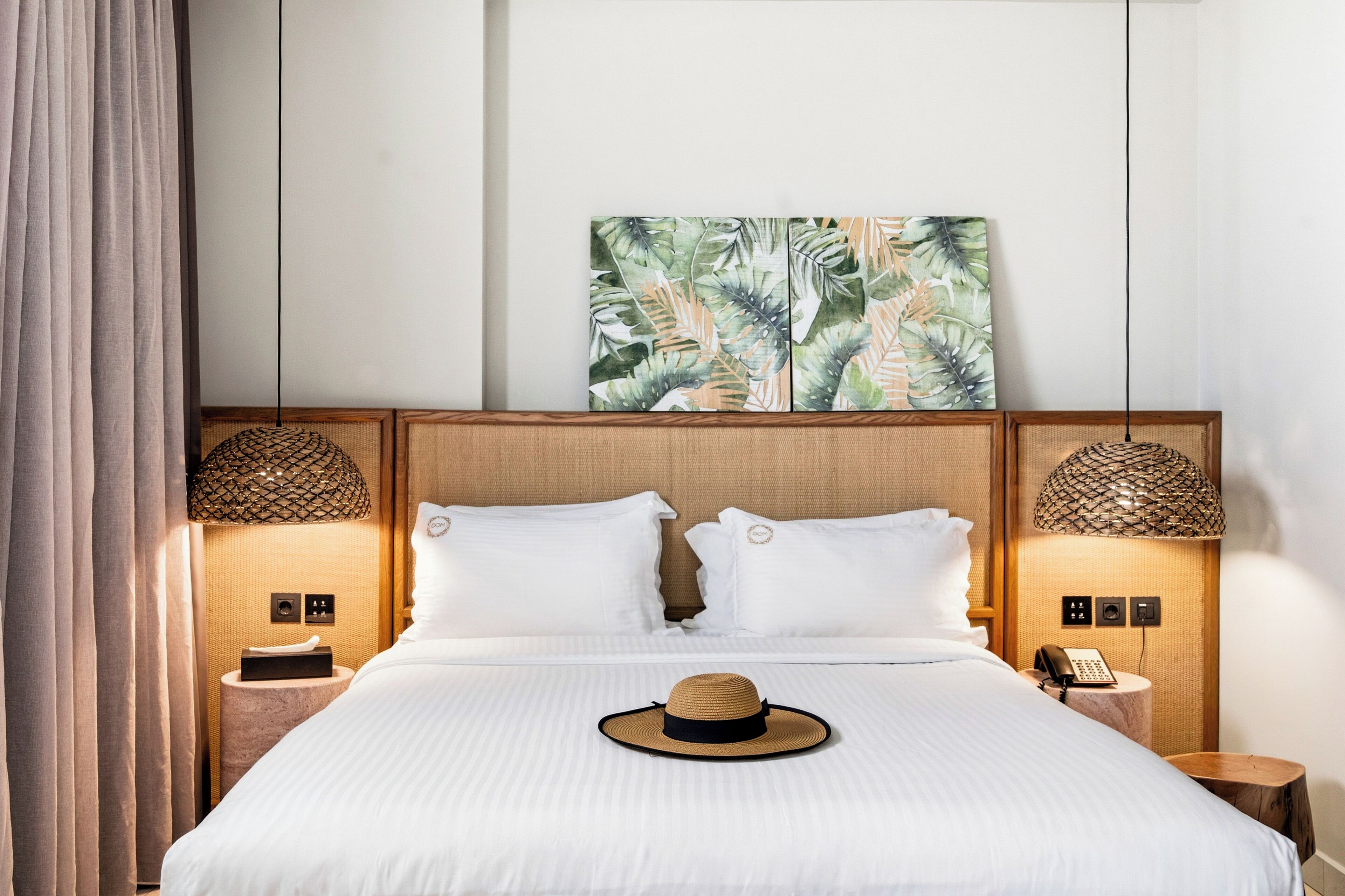 Habitación de hotel con cama, dos mesitas de noche, teléfono montado en la pared y una obra de arte con motivos de hojas tropicales sobre la cama.