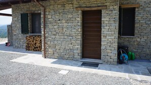 A stone building with a wooden door, a window with shutters, and a firewood stack.