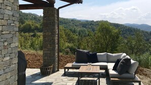 A stone patio with a white sofa and a table, overlooking a forested hillside.