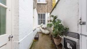 Une ruelle étroite aux murs blancs, avec un sol en béton, un pot de fleurs, un jardinier en forme de main et un portail en métal.