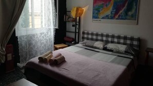 A hotel room with a bed, a window with curtains, a bedside lamp, a bookshelf, and a colorful painting on the wall. - THE GECKO ON THE ROOFS OF GENOA<br>010025-LT-1029 (Genova)