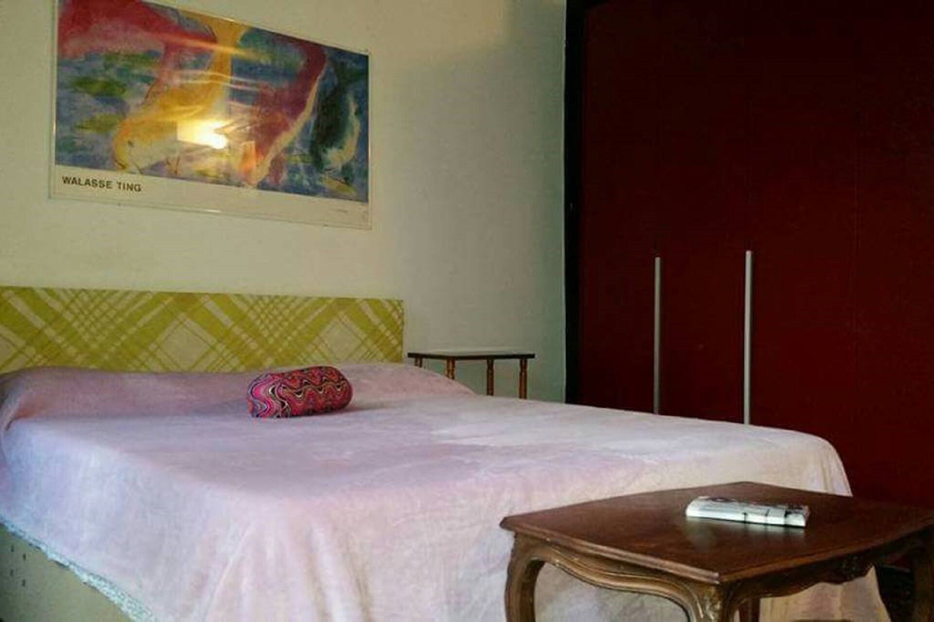 A hotel room with a bed, a bedside table, a colorful abstract painting, and a red wall.