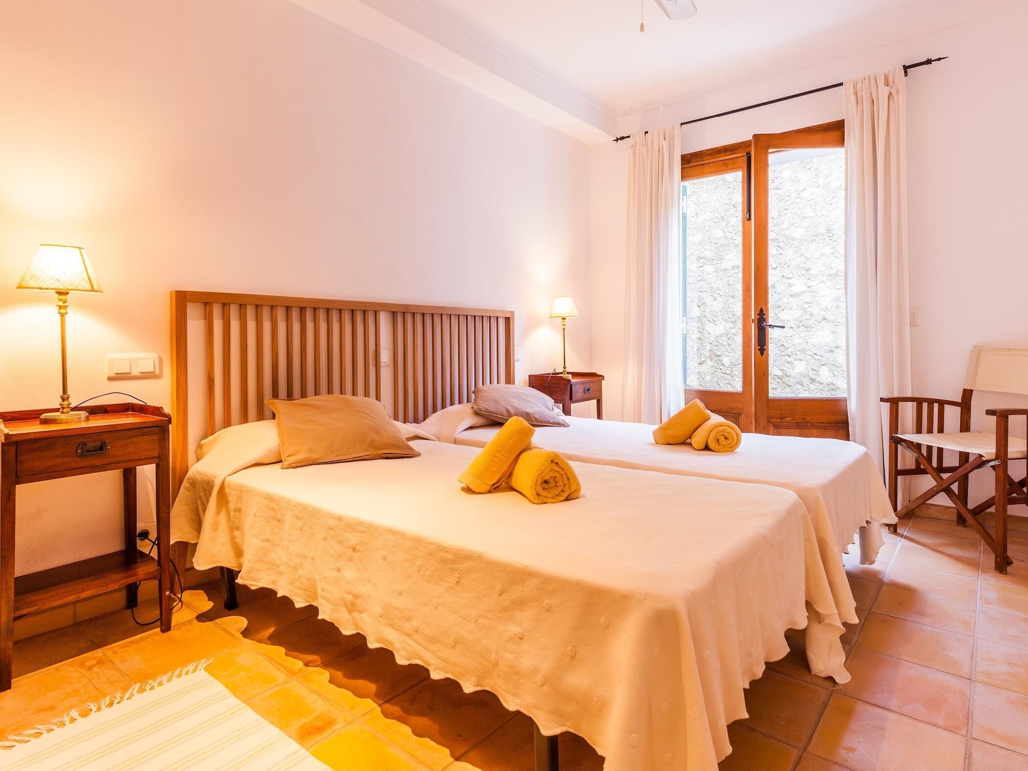 A hotel room with two beds, a wooden headboard, a small desk, a chair, and a window with curtains.
