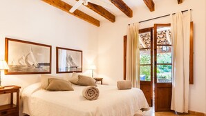 Un dormitorio con cama, almohadas, una toalla enrollada, una mesita de noche, una lámpara, un cuadro con barcos de vela y una ventana con cortinas que ofrece vistas a la vegetación exterior.