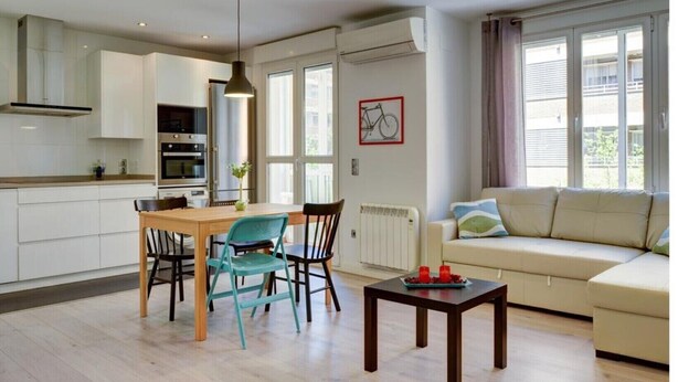 A modern kitchen with white cabinets, a dining table with chairs, a sofa, and a radiator.