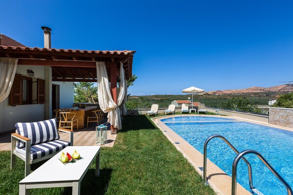 A poolside area with a lounge chair, a table with fruit, and a view of the pool and hills.